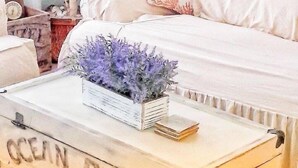 Un cofre de madera de estilo vintage con las palabras "OCEAN PIER" encima, sobre un suelo alfombrado en una habitación con un sofá blanco y un cojín decorativo.
