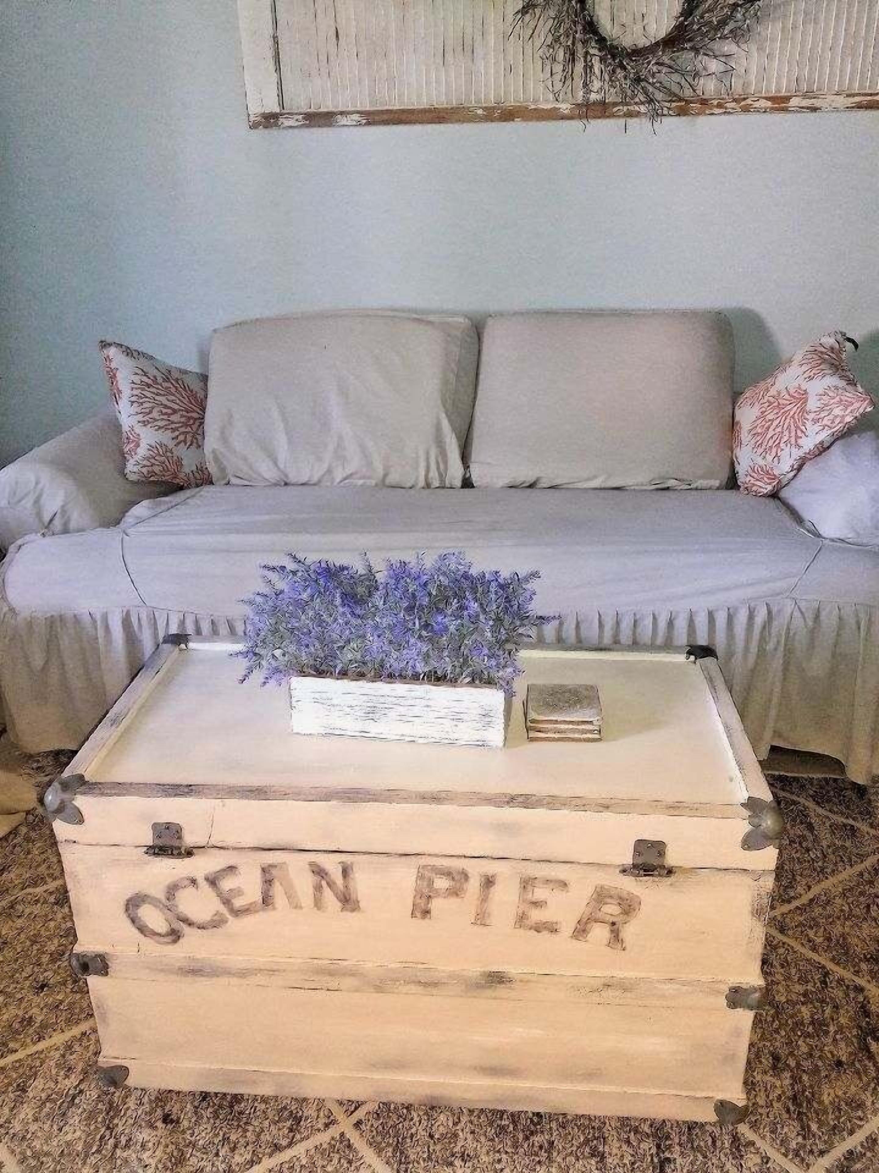 Une pièce avec un canapé de couleur claire, une malle en bois portant l’inscription « OCEAN PIER », et une petite plante posée sur la malle.