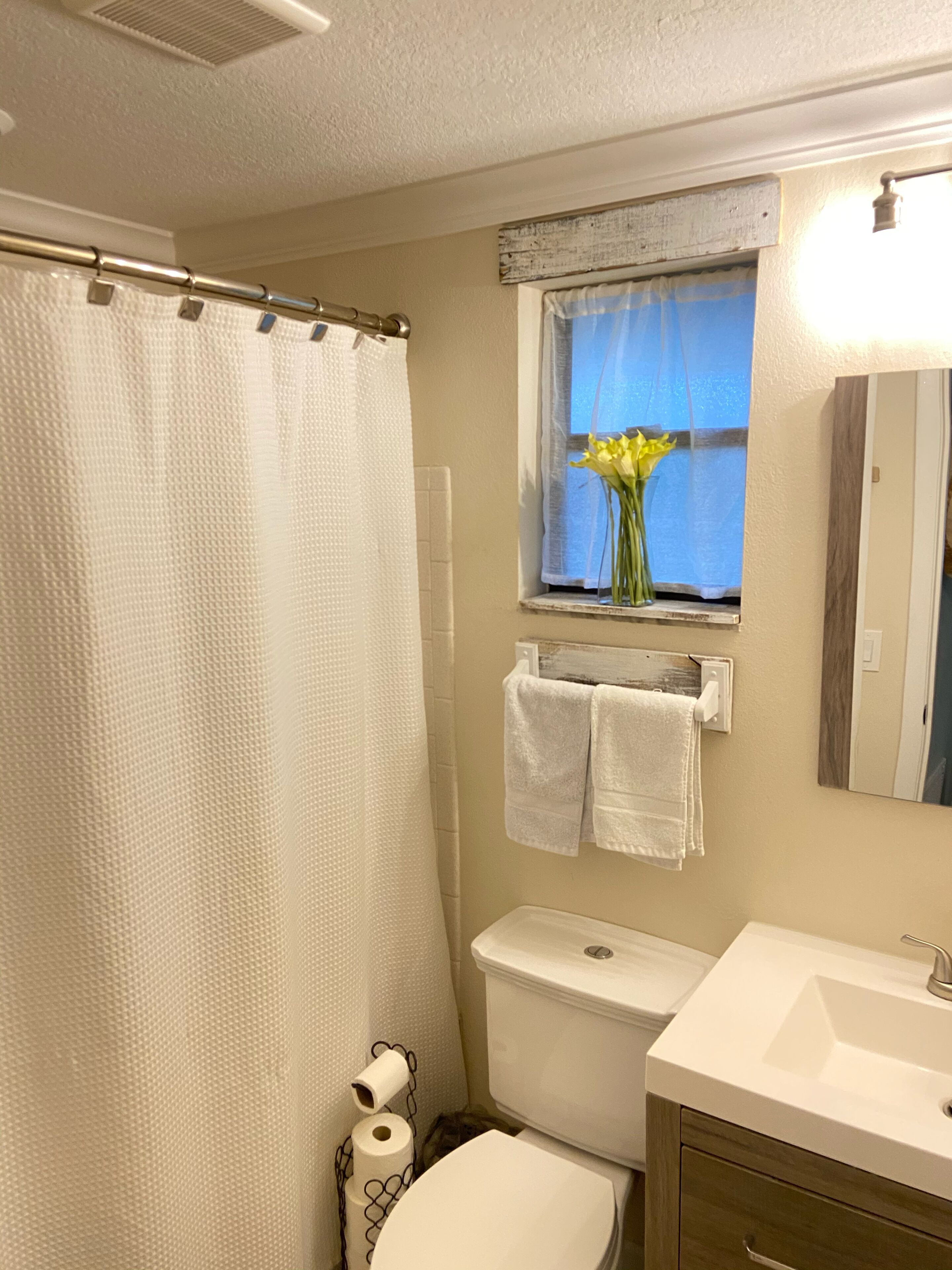 Un baño con cortina de ducha, inodoro, lavabo, una ventana con un florero y un perchero para toallas.