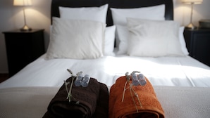 Una cama bien tendida con sábanas blancas y dos toallas enrolladas, una marrón y otra naranja, cada una con un pequeño adorno.