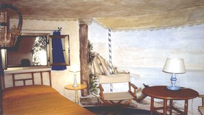Une pièce avec un lit en bois, une petite table avec une lampe, une chaise, une fenêtre avec un rideau et une peinture murale.