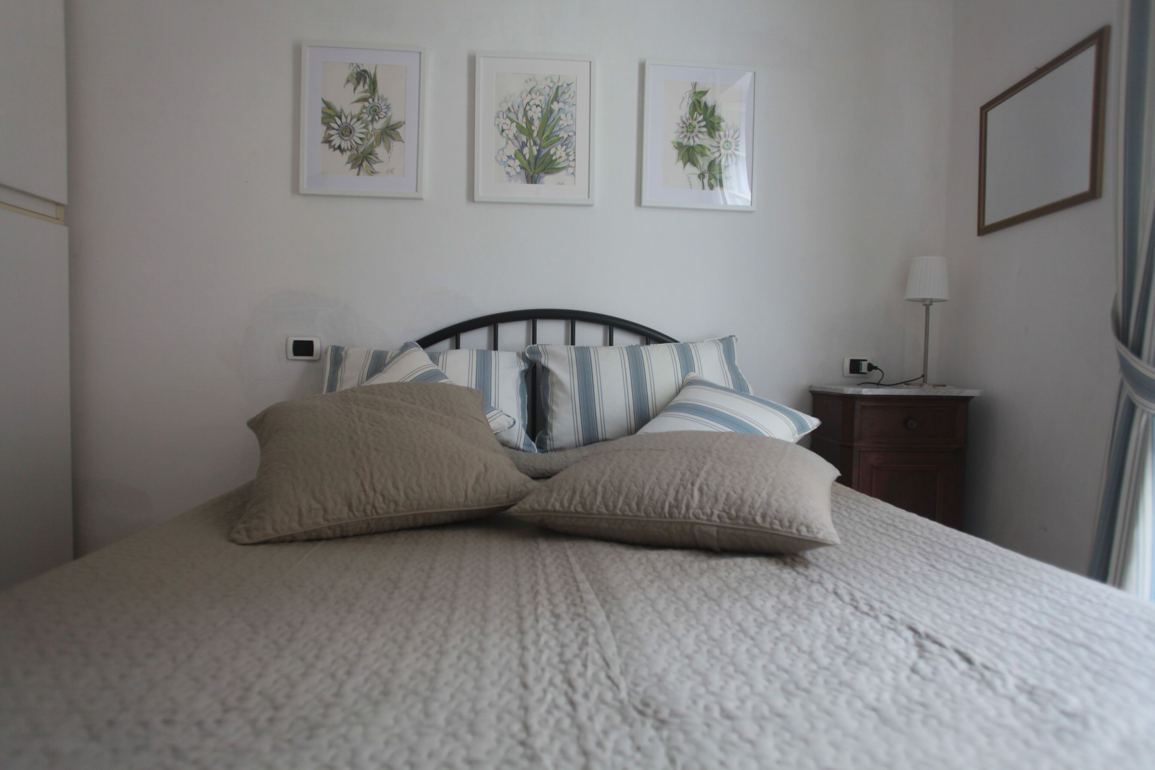 A bed with a striped headboard, two pillows, and a bedside table with a lamp. There are three framed botanical prints on the wall above the bed.