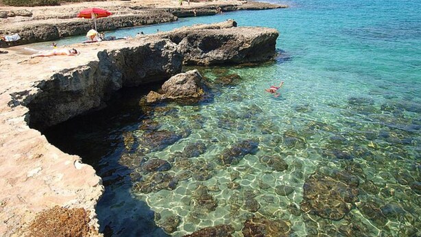 Una playa rocosa con agua cristalina, una sombrilla roja y una persona nadando.