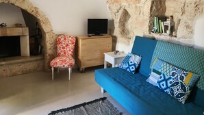 Una habitación acogedora con pared de piedra, un sofá azul con cojines estampados, un sillón rojo con estampado floral, un aparador de madera y un televisor sobre una mesita.