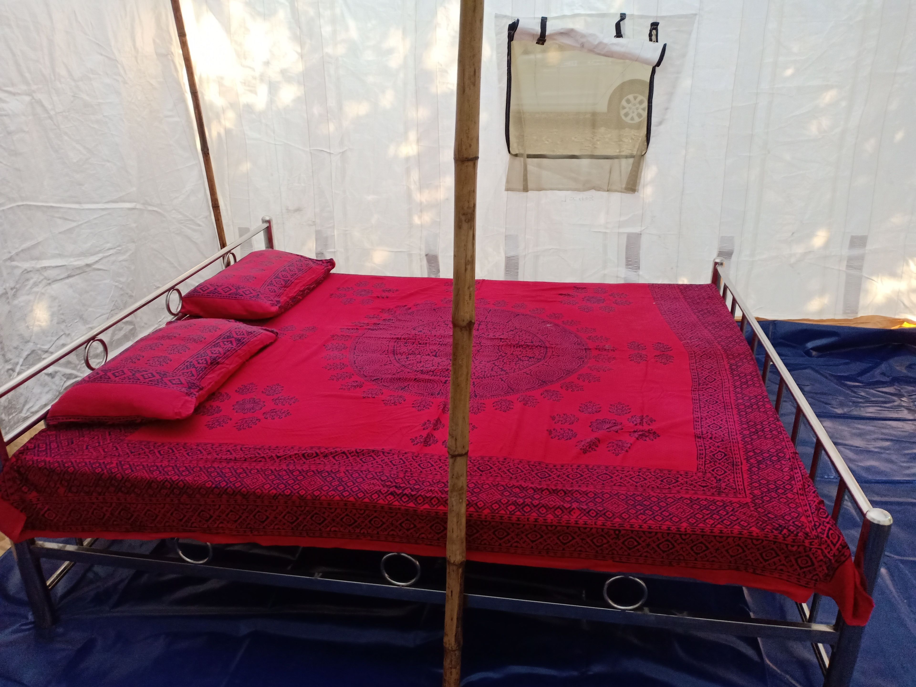 Una cama individual con una colcha roja estampada y dos almohadas, estructura de metal y una ventana con cortina translúcida.
