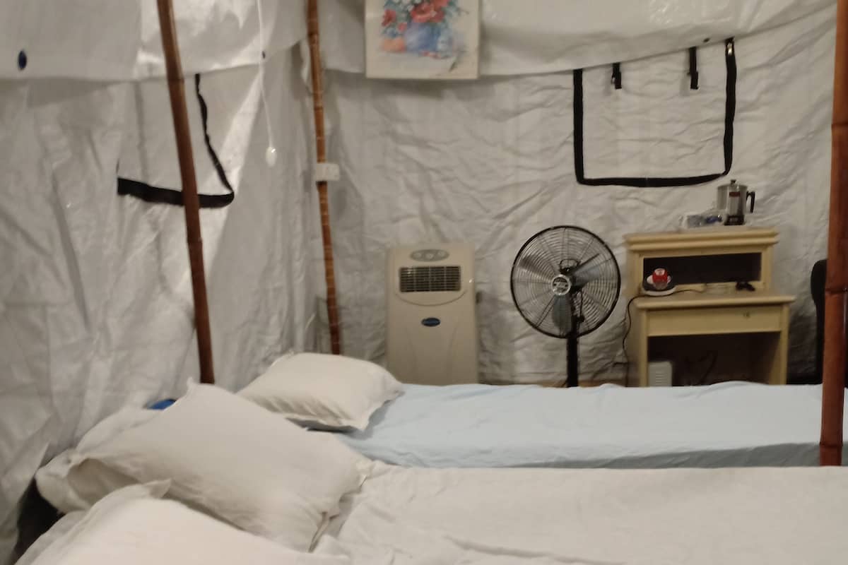 Una habitación con dos camas, un ventilador, un calentador y una pequeña mesa con un hervidor.