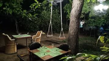 An outdoor dining area with tables and chairs, surrounded by trees and a hanging light fixture.