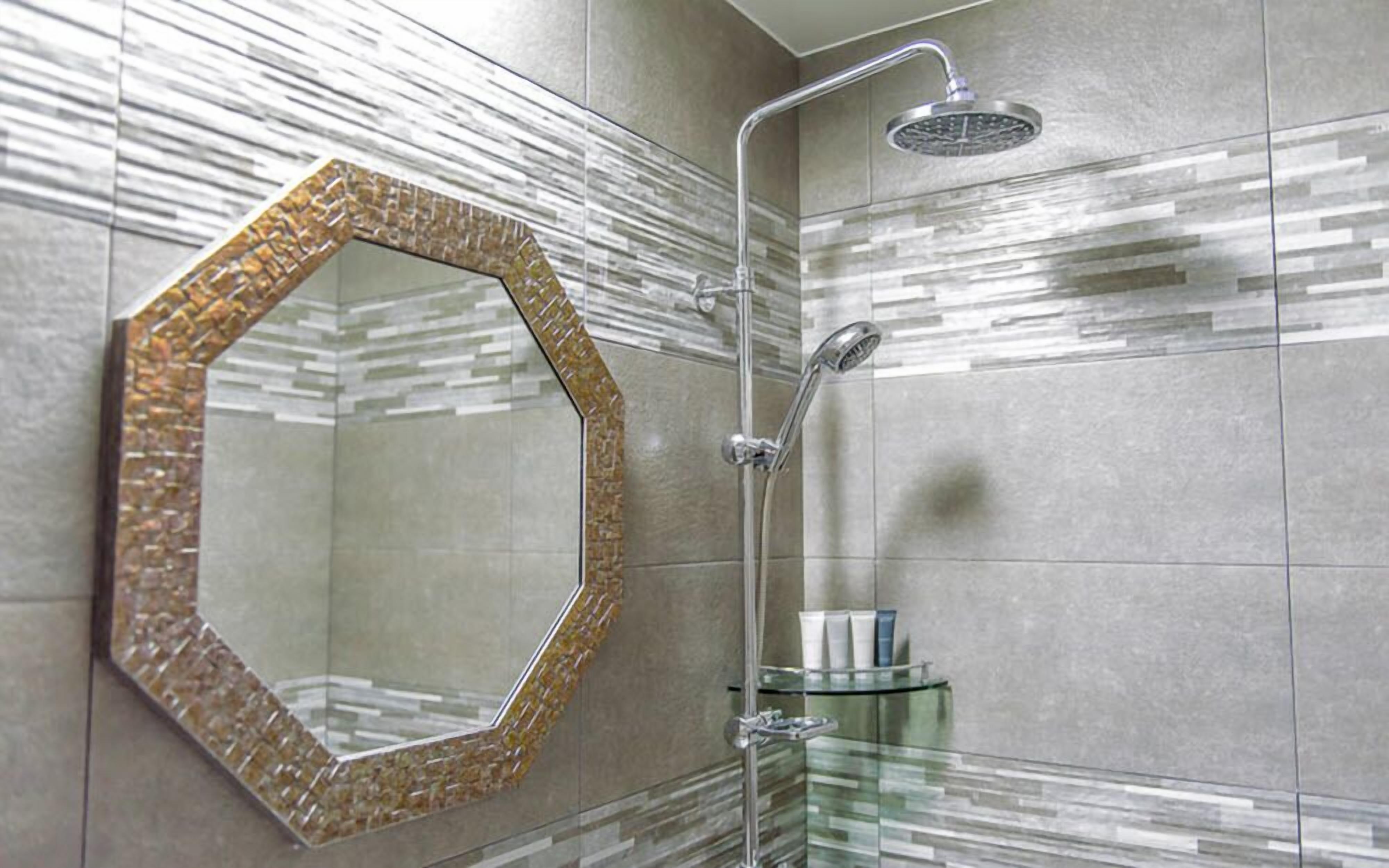 A bathroom with a hexagonal mirror, a glass shower enclosure, and a glass shelf with toiletries.