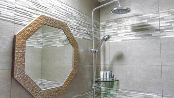 A bathroom with a hexagonal mirror, a glass shower enclosure, and a glass shelf with toiletries.