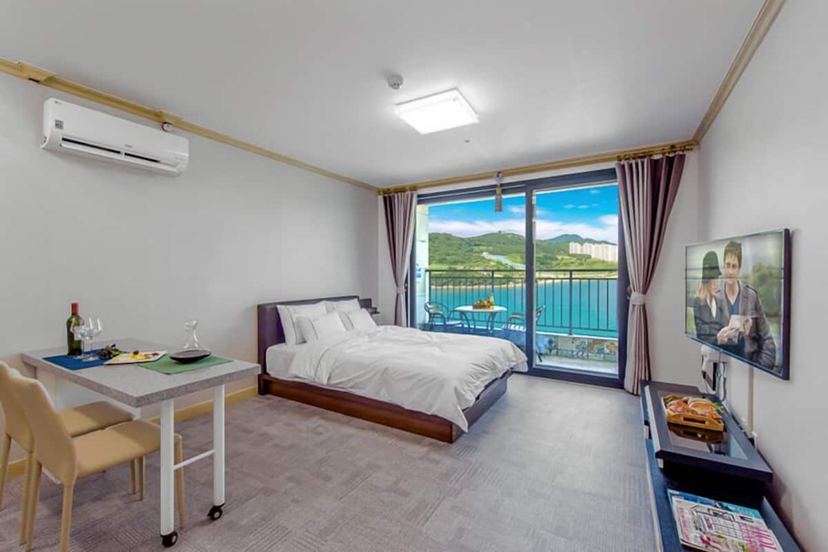 A hotel room with a bed, a desk with a chair, a TV, and a balcony with a view of the sea.
