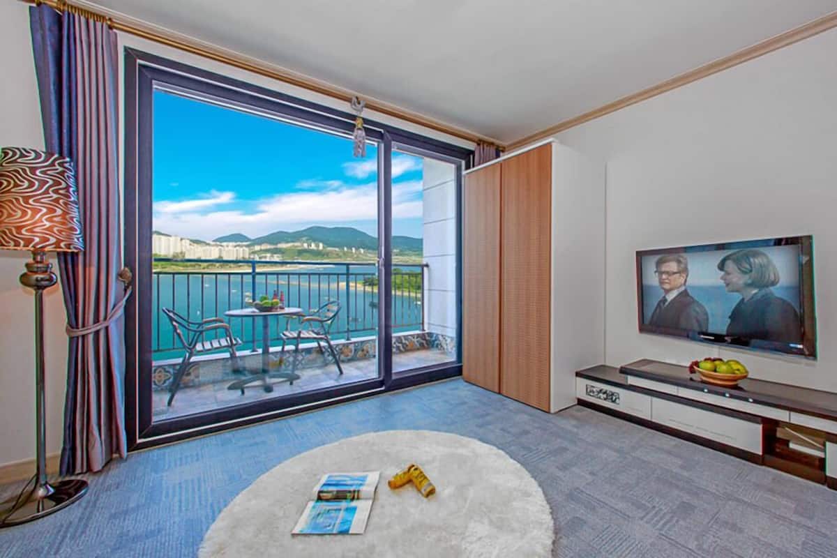 A modern hotel room with a large sliding glass door leading to a balcony with a view of the sea and mountains. There is a TV on the wall, a small round rug, and a table with chairs.