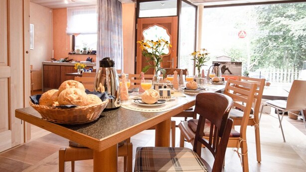 A breakfast setting with a wooden table, chairs, and a basket of bread. - Gästehaus Im Tal 18 (Hellenthal)