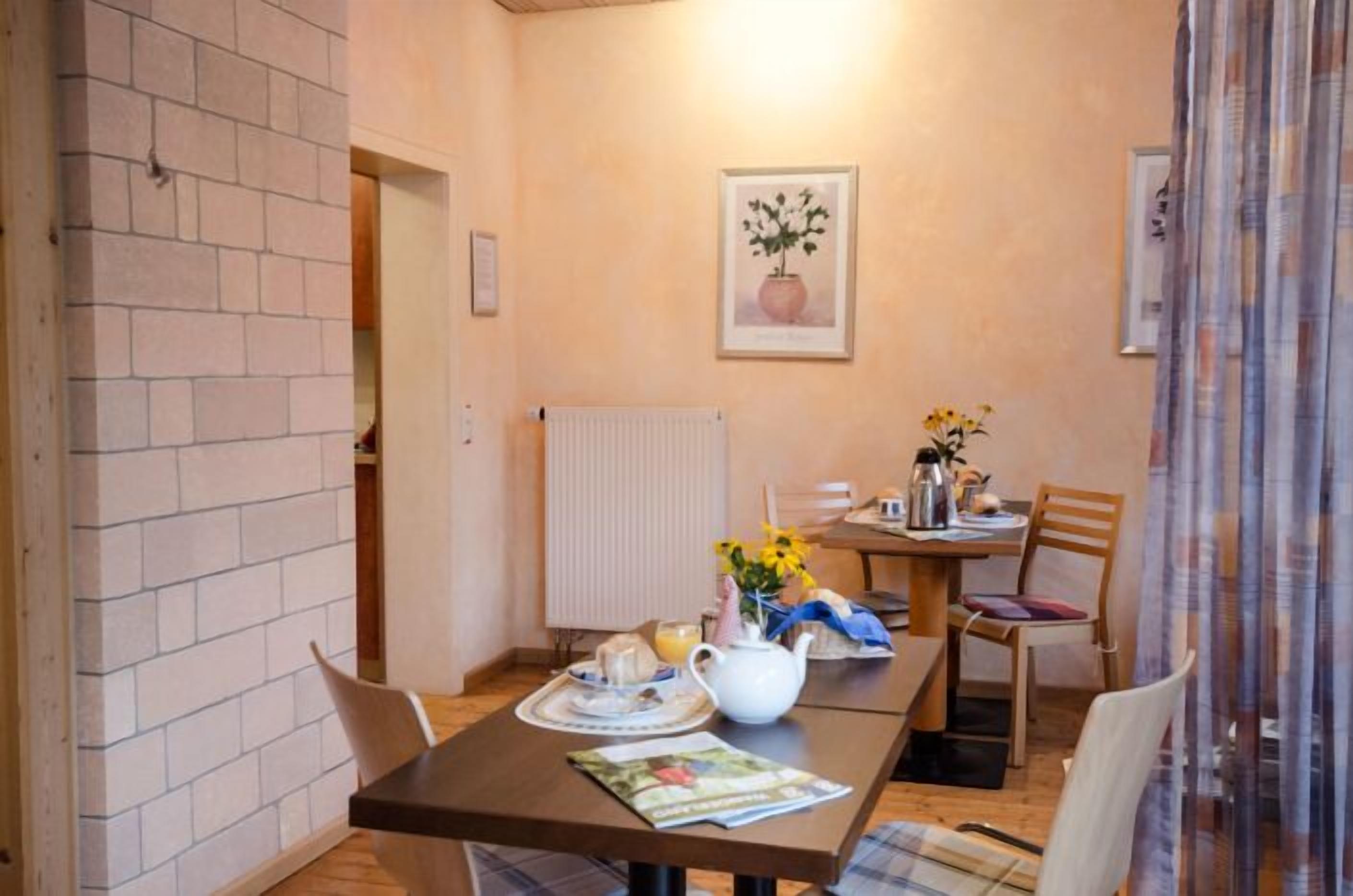 A dining area with a table set for two, a radiator, a framed picture on the wall, and a window with curtains.
