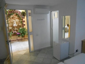 Une pièce comprenant un lit, une petite armoire blanche, un miroir et une porte donnant sur un jardin agrémenté d’une statue et de plantes.