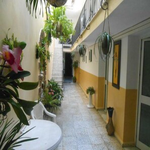 Un couloir étroit au sol carrelé, avec des chaises blanches et des plantes en pot.