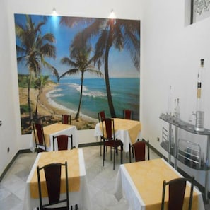 Un restaurant décoré d’une fresque représentant une plage, avec des tables recouvertes de nappes jaunes et un bar où sont exposées des bouteilles.