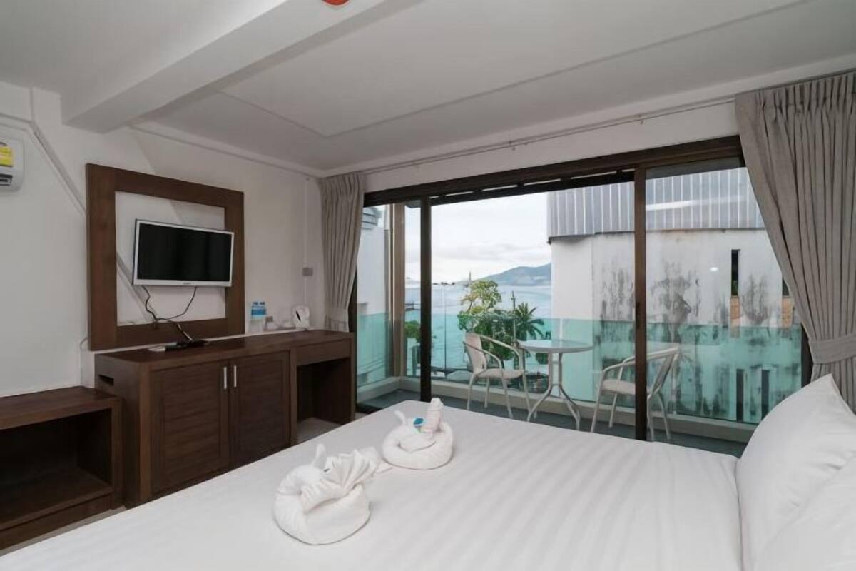 A hotel room with a bed, a TV, a desk, a balcony with a table and chairs, and a view of the sea.