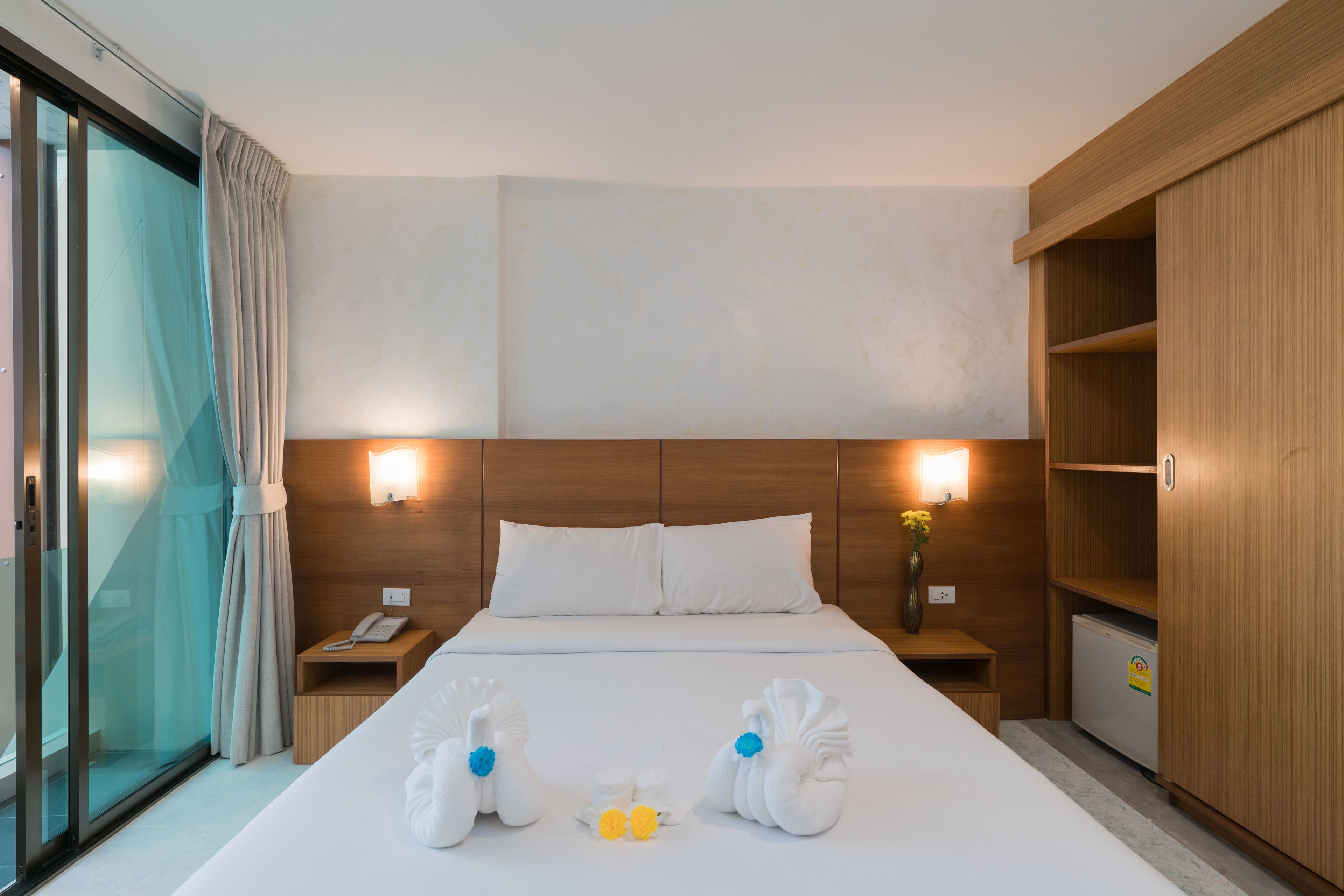 Habitación de hotel con una cama grande, dos mesitas de noche, un armario de madera y un ventanal corredizo.