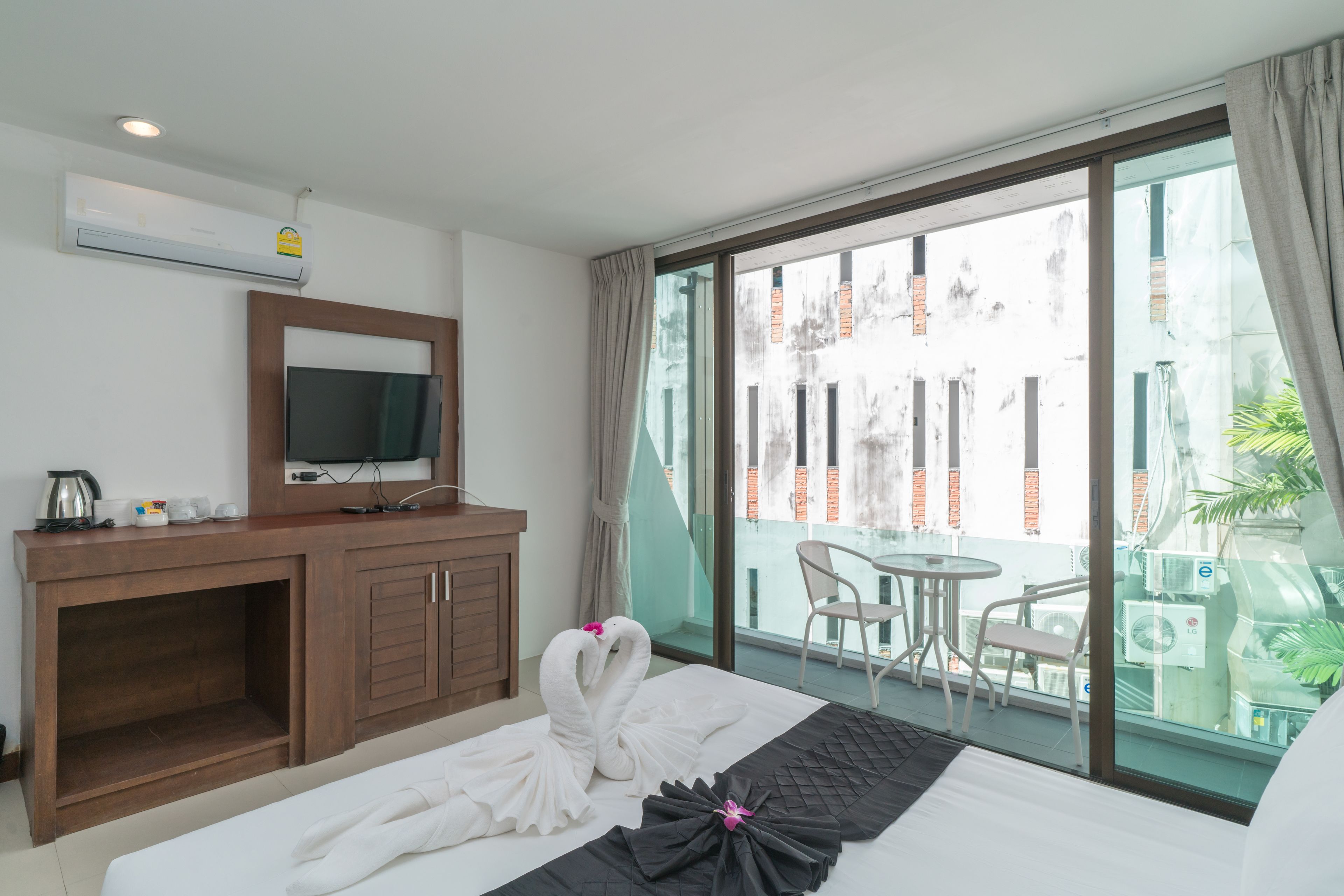 Une chambre d’hôtel avec un lit, un téléviseur posé sur une commode en bois, un balcon avec une table et des chaises, et une vue sur les bâtiments environnants.