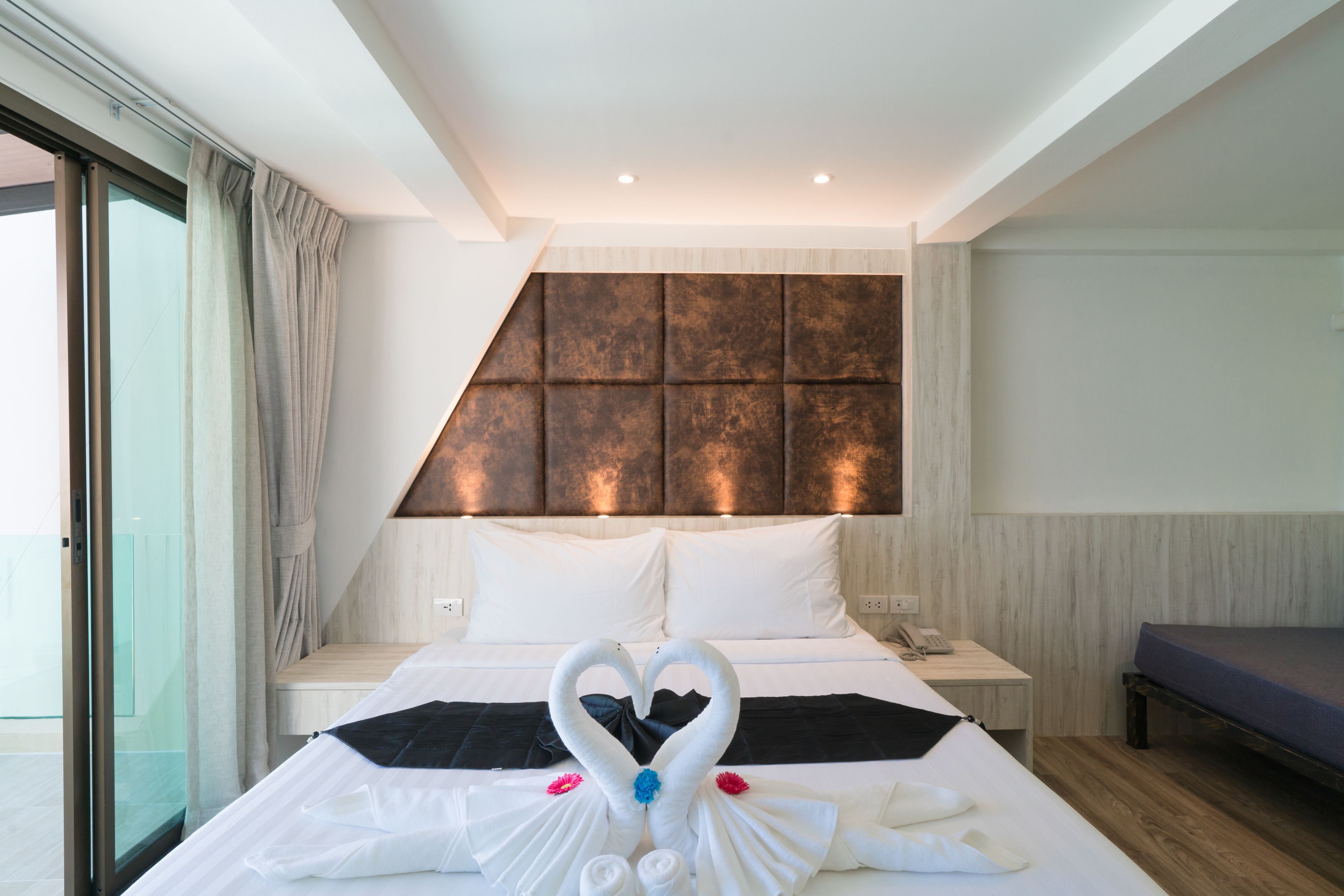 A modern hotel room with a large bed, a headboard with a textured panel, a bedside table with a phone, and a bench by the window.