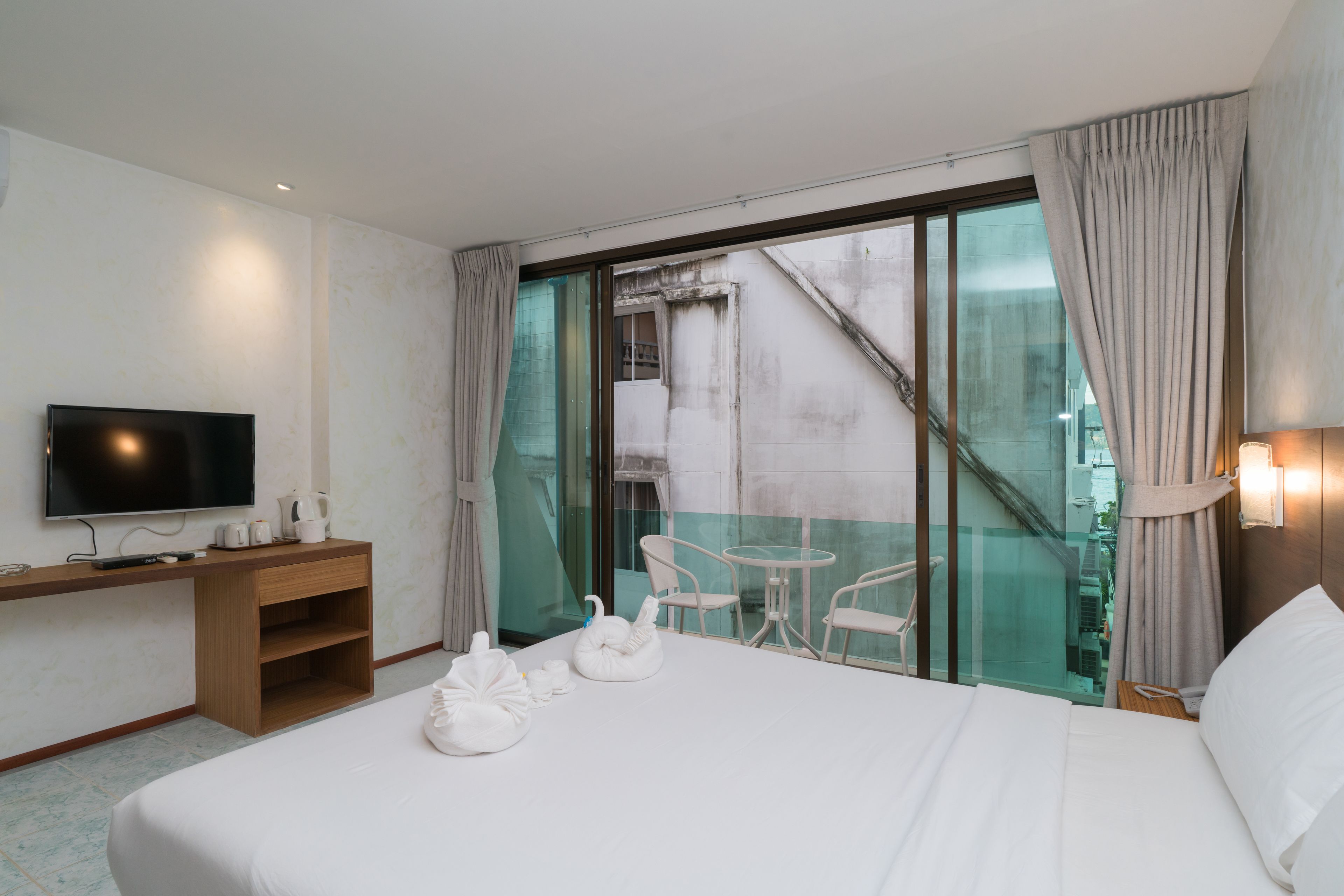 A hotel room with a bed, a desk with a TV, a balcony with a glass door, and a view of the city.