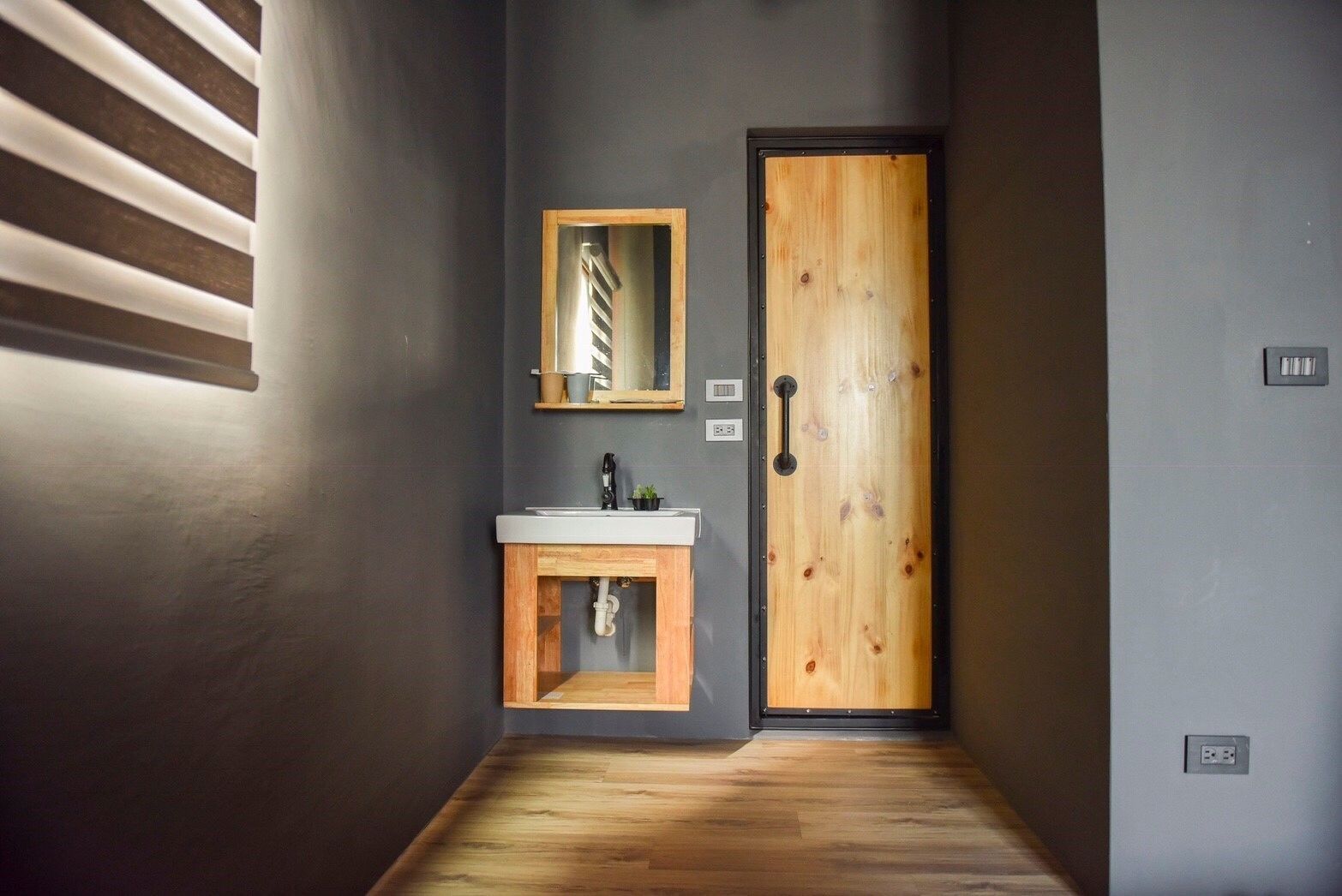 A hallway with a wooden door, a small sink with a mirror, and a light fixture on the wall.