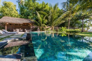 A clear swimming pool with lounge chairs and a thatched-roof structure. - The Beach Villa (Tanjung)
