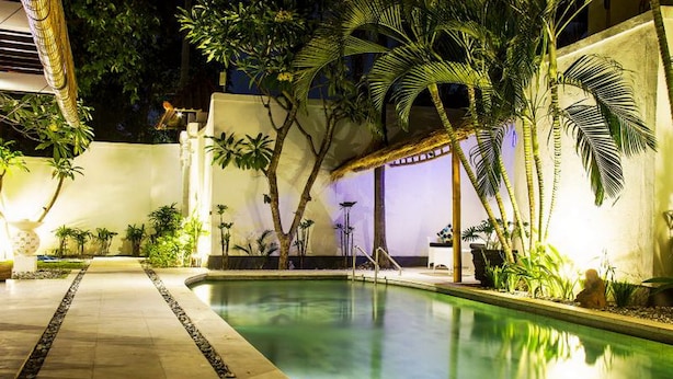 Un área de piscina nocturna con piscina cristalina, palmeras y un camino que lleva a un área de descanso cubierta.