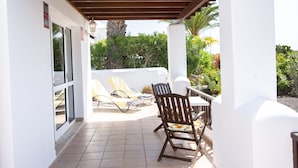 A covered outdoor patio with a table and two chairs, a door, and a view of the surrounding greenery.