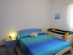 A bedroom with a bed, a nightstand, a lamp, a window with curtains, and a wall-mounted shelf. - Homely apartment with private garden (Bibione)