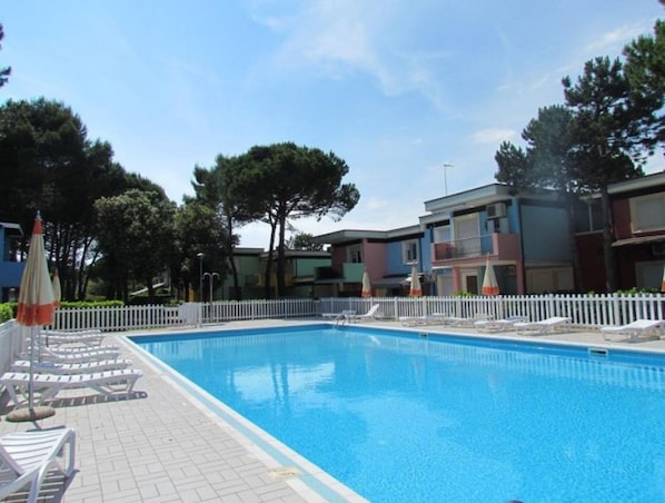 Una piscina con sillas de descanso y sombrillas, rodeada por una valla blanca y árboles.