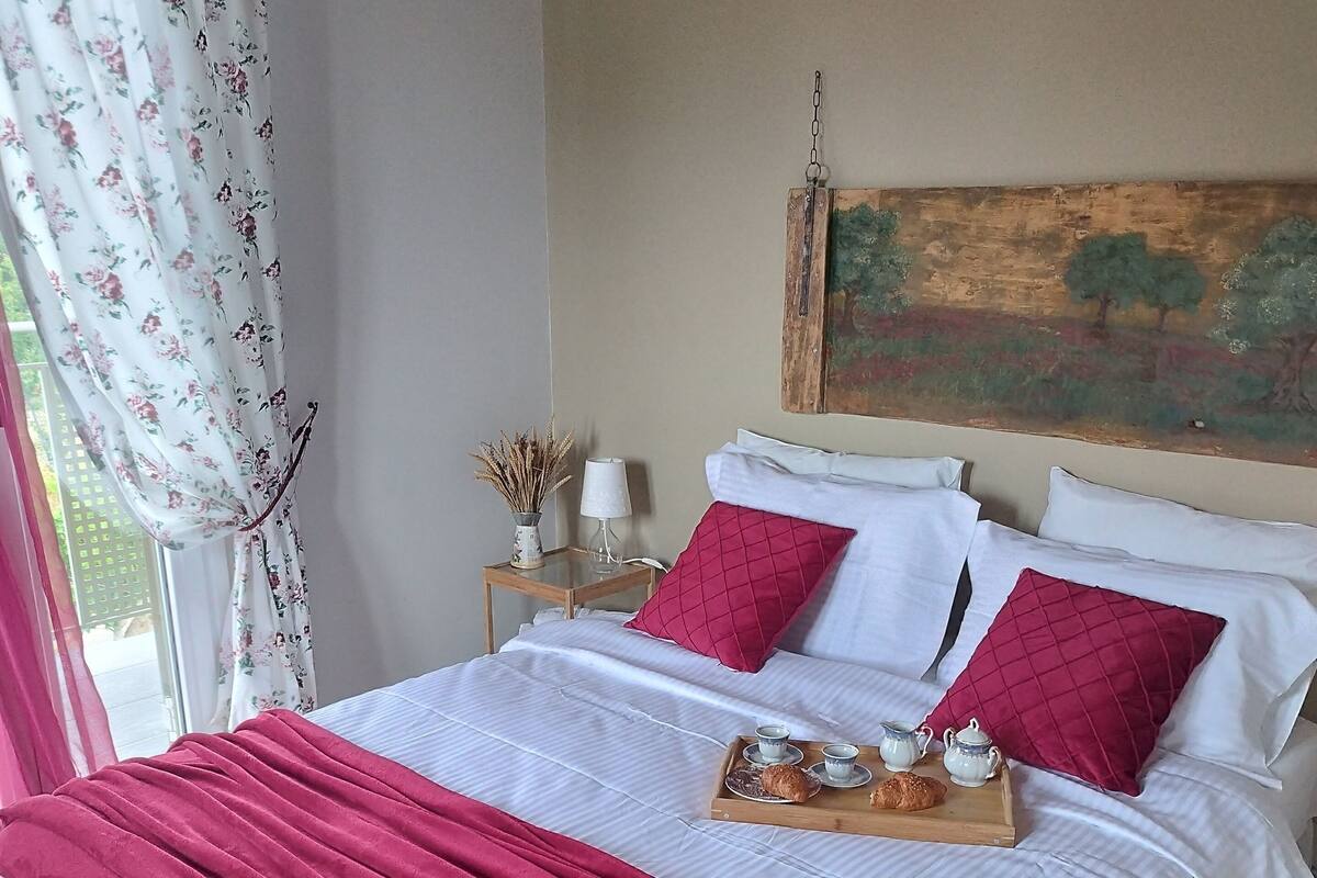 Un lit bien fait, avec du linge de lit blanc et des touches de rouge, un plateau avec des éléments de petit-déjeuner et un tableau accroché au mur.