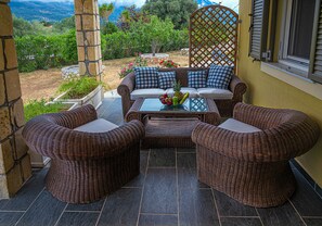 A patio with wicker furniture, a glass table with fruit, and a view of the garden.
