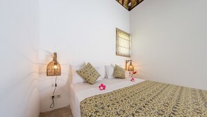 A bedroom with a patterned bedspread, two wall-mounted lamps, and a small window with a patterned curtain. - Jambu Villa Lombok (Tanjung)