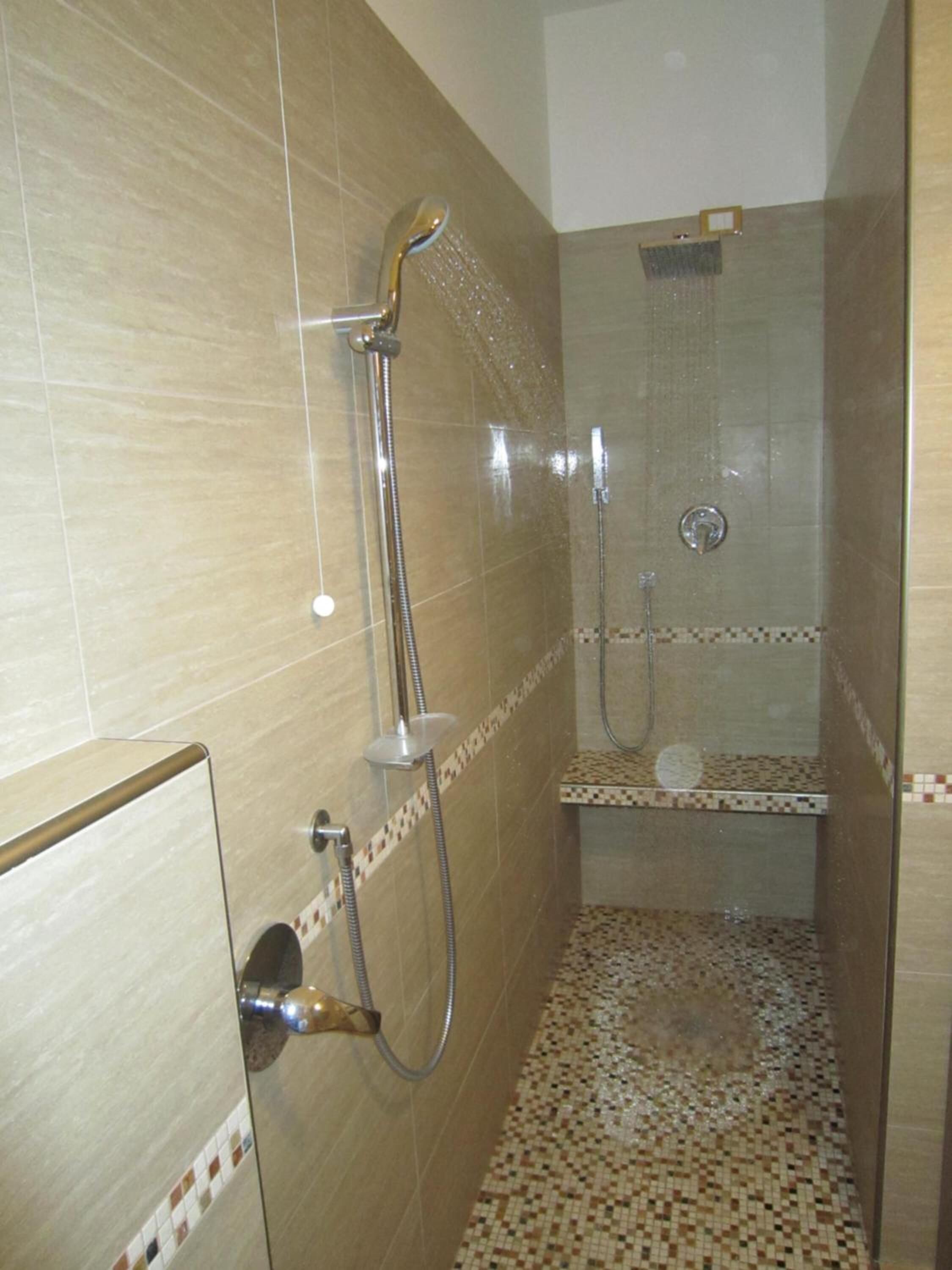 A shower area with a handheld showerhead and a wall-mounted showerhead, tiled walls, and a tiled floor.