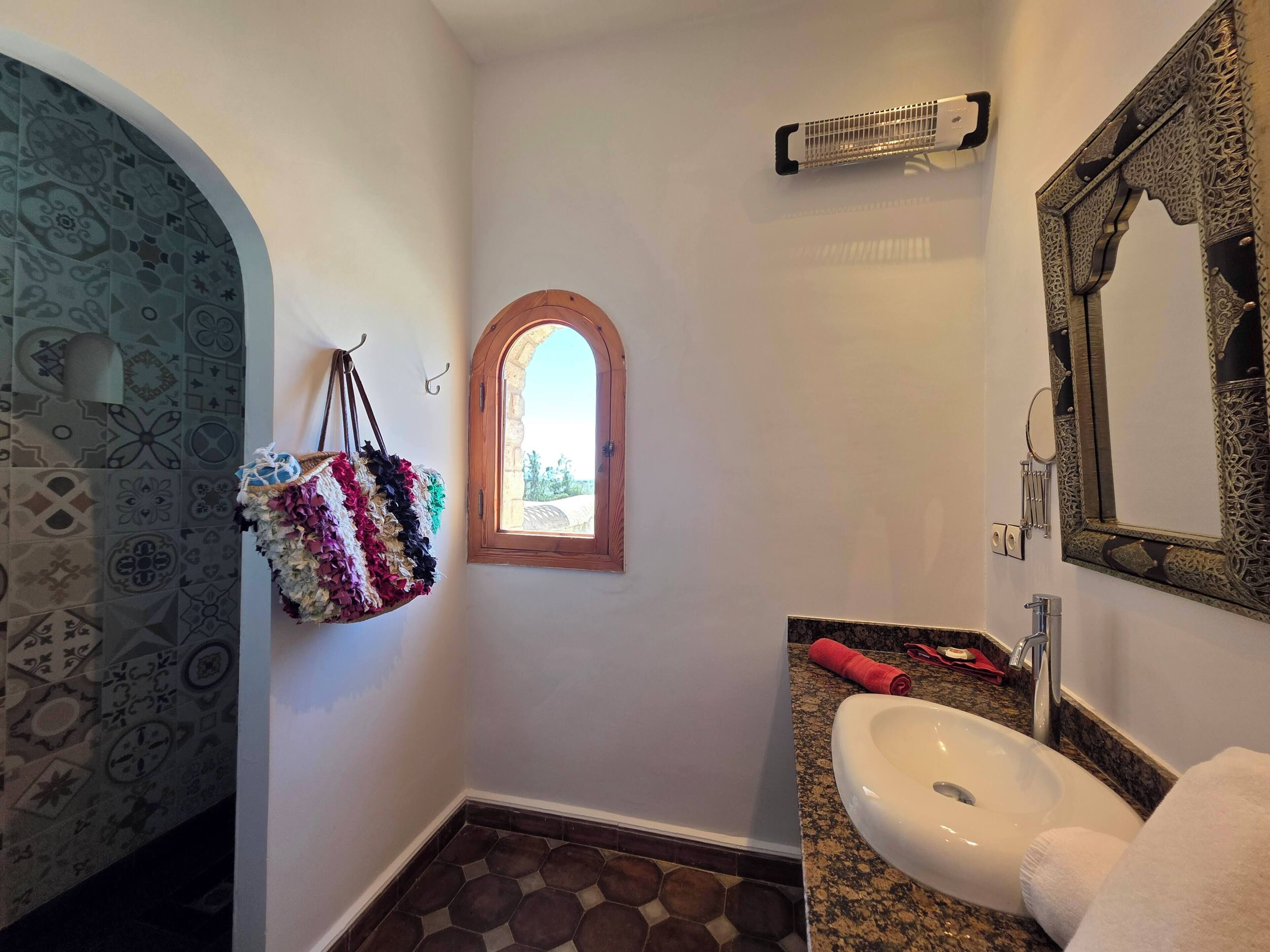 Une salle de bain avec un mur recouvert de carreaux à motifs, une petite fenêtre, un lavabo avec une serviette et un miroir décoratif.
