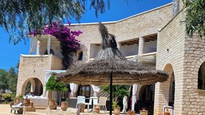 Un edificio de piedra con piscina, sillones de descanso y un toldo de paja.