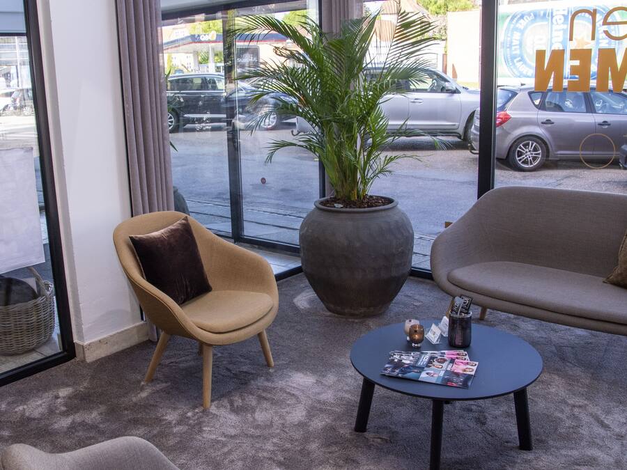 Un área de recepción con un sofá, un sillón y una mesita con revistas. Hay una planta grande en maceta y una cesta de mimbre cerca de la entrada.