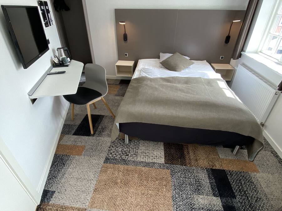 Una habitación de hotel moderna con una cama, mesitas de noche, un escritorio con una silla, un televisor y una ventana.