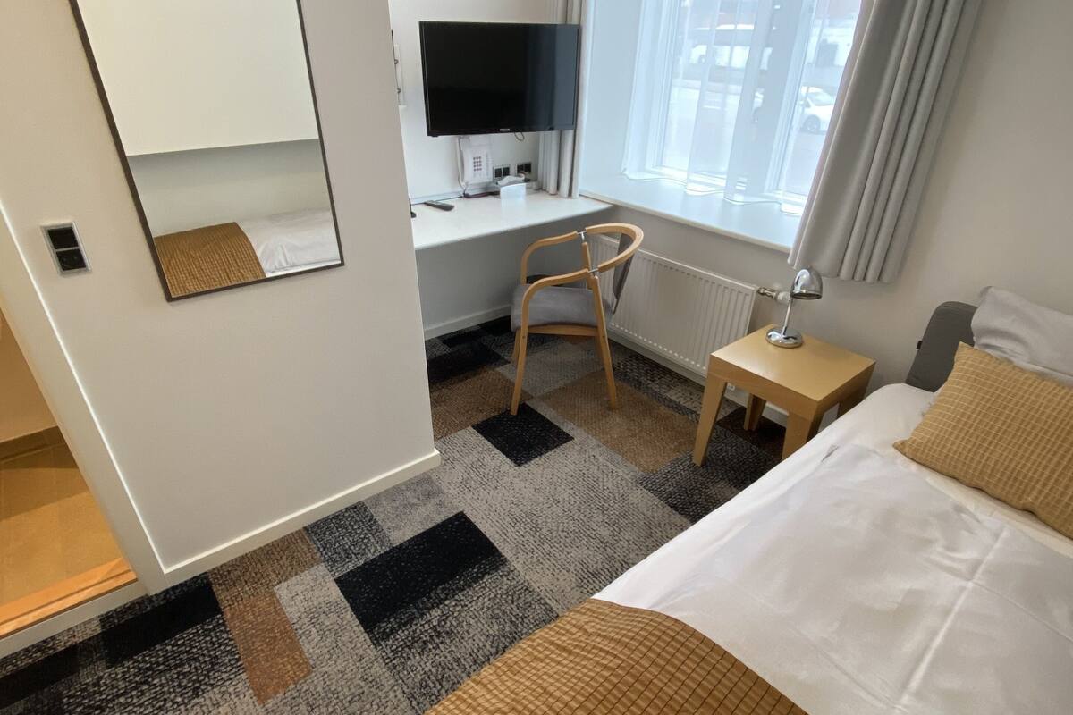 Habitación de hotel con cama, escritorio con computadora, silla, ventana con cortinas y espejo.
