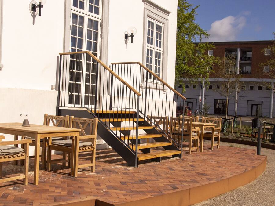 An outdoor seating area with wooden tables and chairs, a staircase leading to a building entrance, and a clear view of the surrounding architecture.