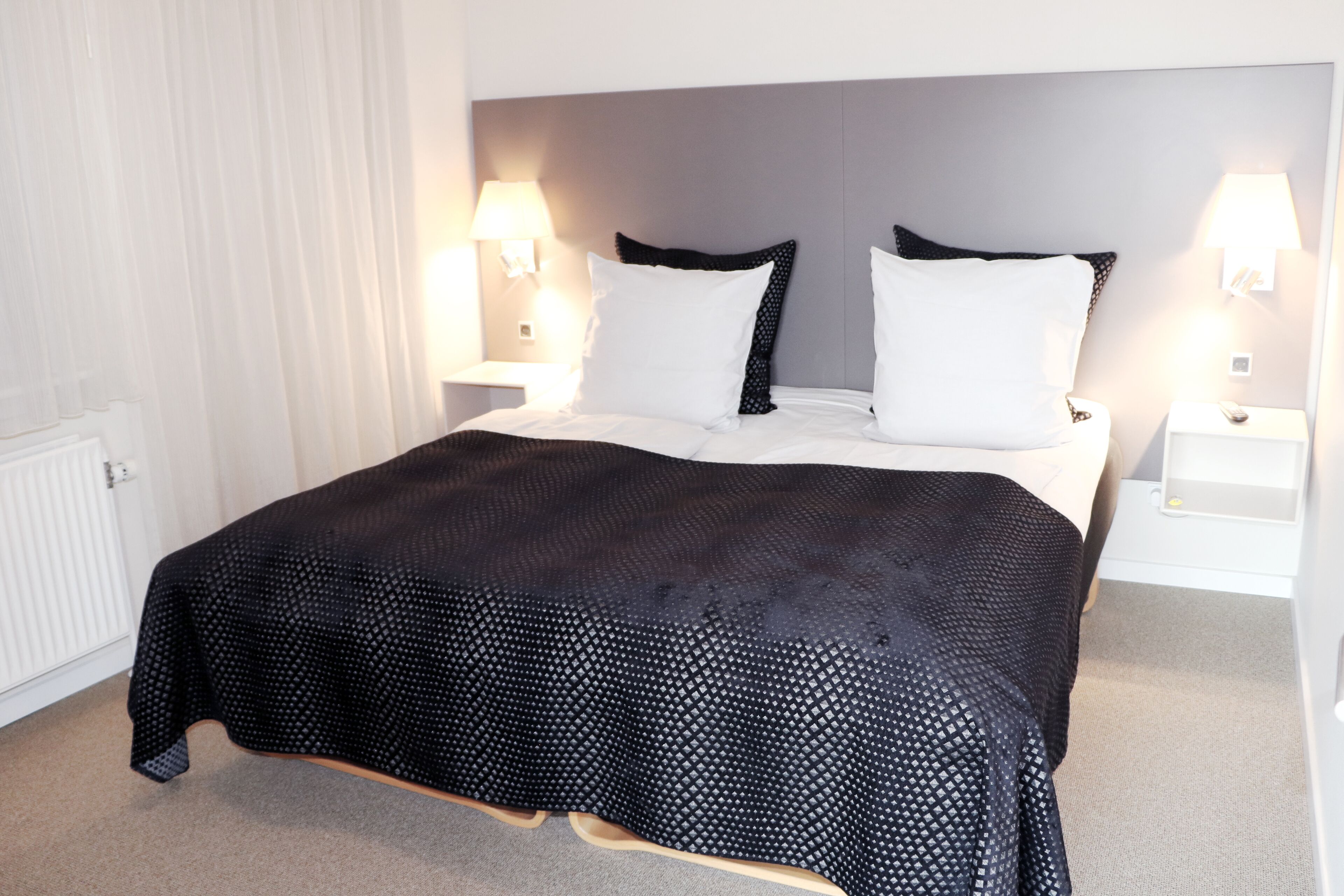Ein ordentlich bezogenes Bett mit einem schwarz-weiß gemusterten Überwurf, zwei weißen Kissen und je eine Nachttischlampe auf beiden Seiten.