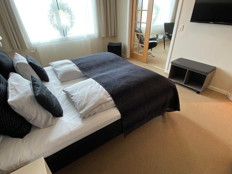 Una cama bien hecha con ropa de cama blanca y negra, una mesita de noche pequeña, un control remoto y un televisor en la pared.