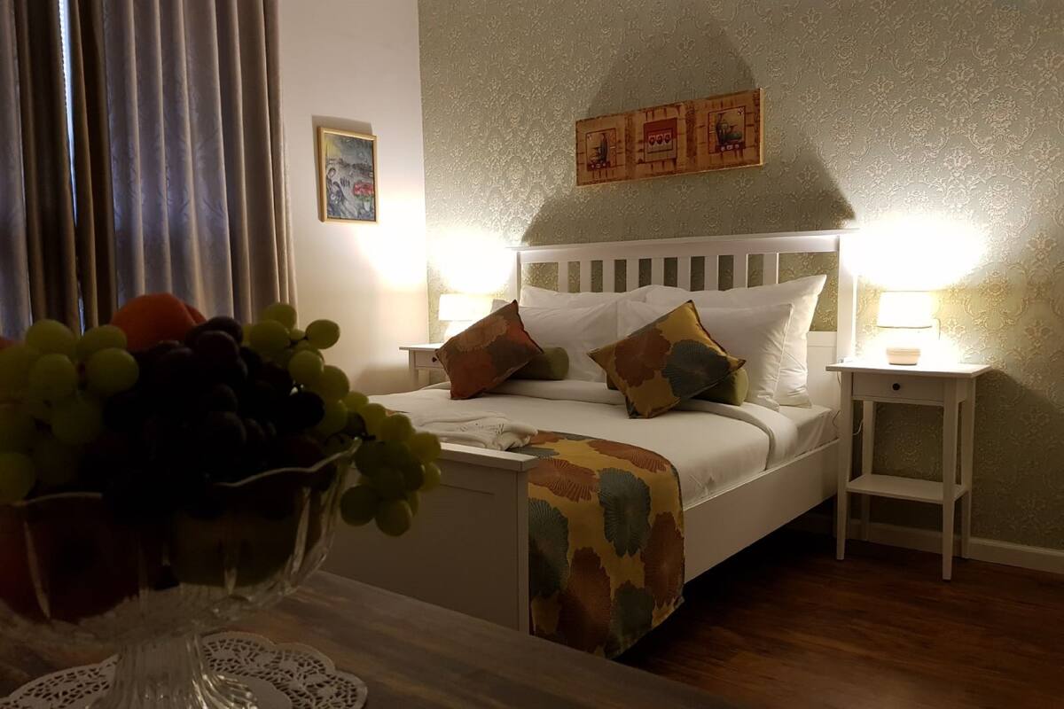 Una cama bien hecha con una manta estampada, dos mesitas de noche con lámparas, un jarrón con uvas sobre una mesa y una pared con un panel decorativo.