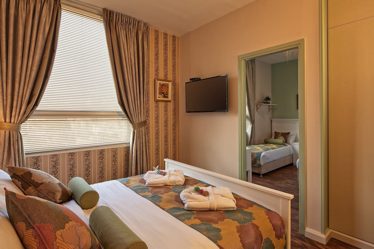 Una habitación de hotel con cama, cortinas, televisor, espejo y vista hacia otra habitación.