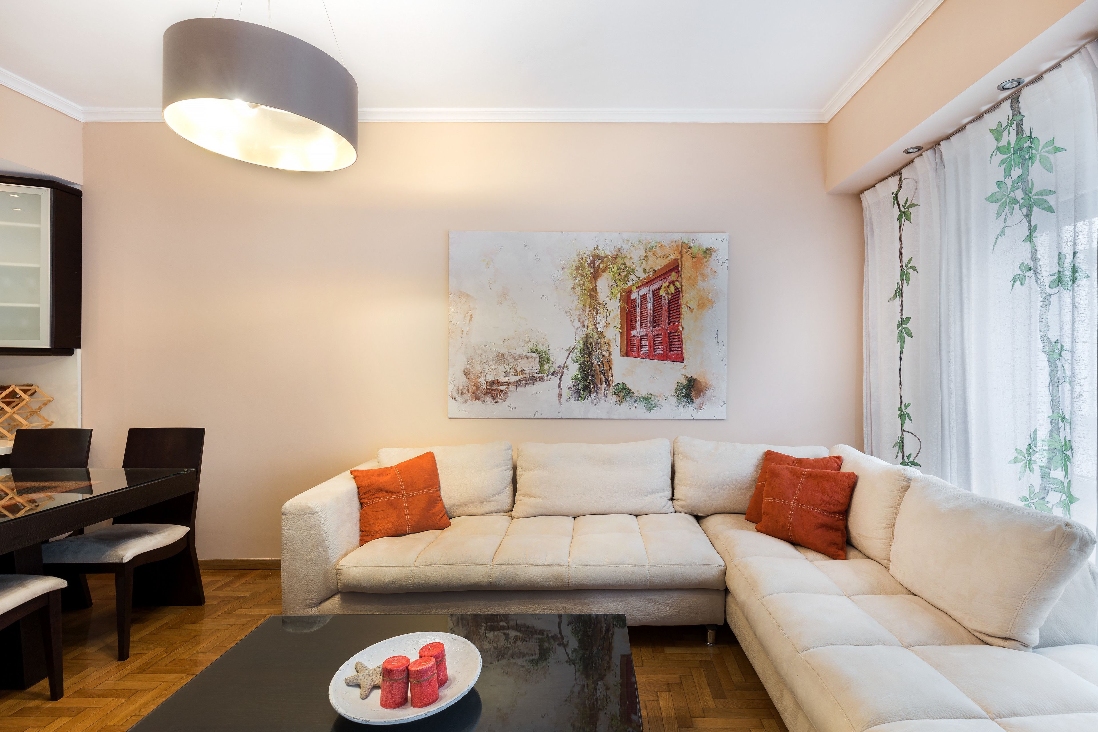 Een woonkamer met een bank, een eettafel en stoelen. Er hangt een schilderij aan de muur en er is een raam met gordijnen.