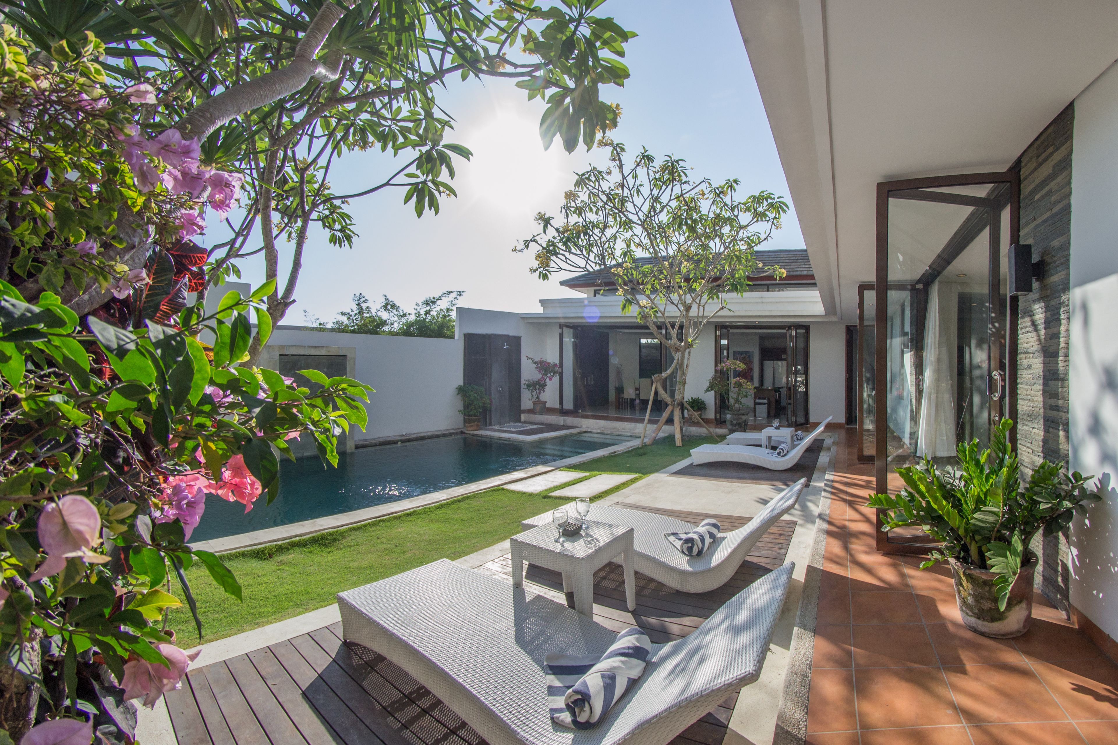 A modern outdoor pool area with lounge chairs and a small table, surrounded by greenery and a clear sky.