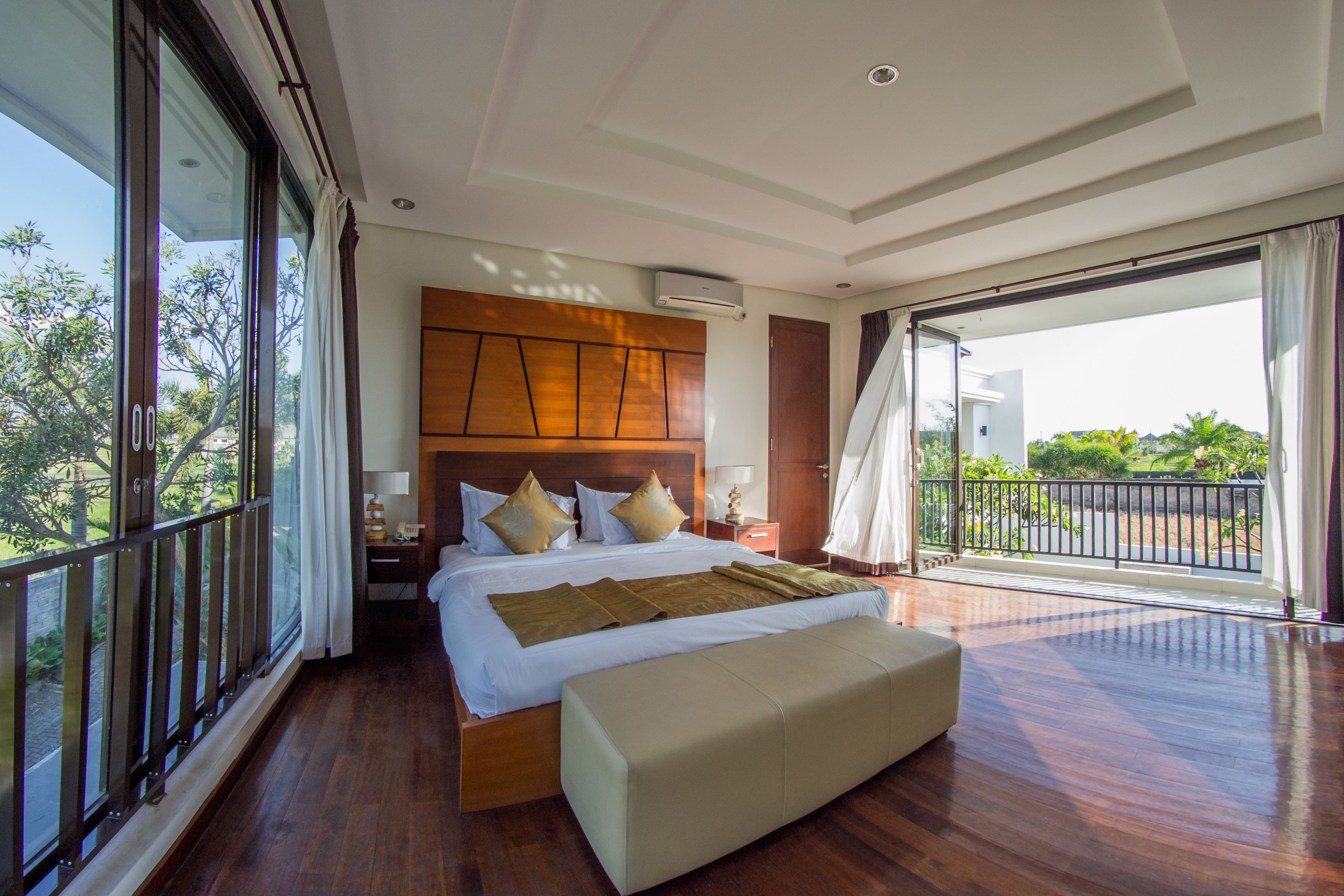 A spacious bedroom with a large bed, a bench, and a balcony with a view of greenery.
