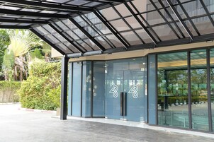 A modern hotel entrance with a glass facade, a metal roof structure, and a well-maintained garden area.