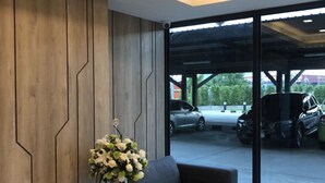 A modern interior with a dark sofa, a small table with a vase of flowers, and a large window overlooking a parking area. - The 28th Hotel (Kanchanaburi)